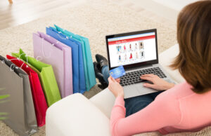 What makes the people tend towards shopping? A girl sitting on sofa and hold the laptop, credit card and shopping near to sofa