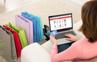 What makes the people tend towards shopping? A girl sitting on sofa and hold the laptop, credit card and shopping near to sofa